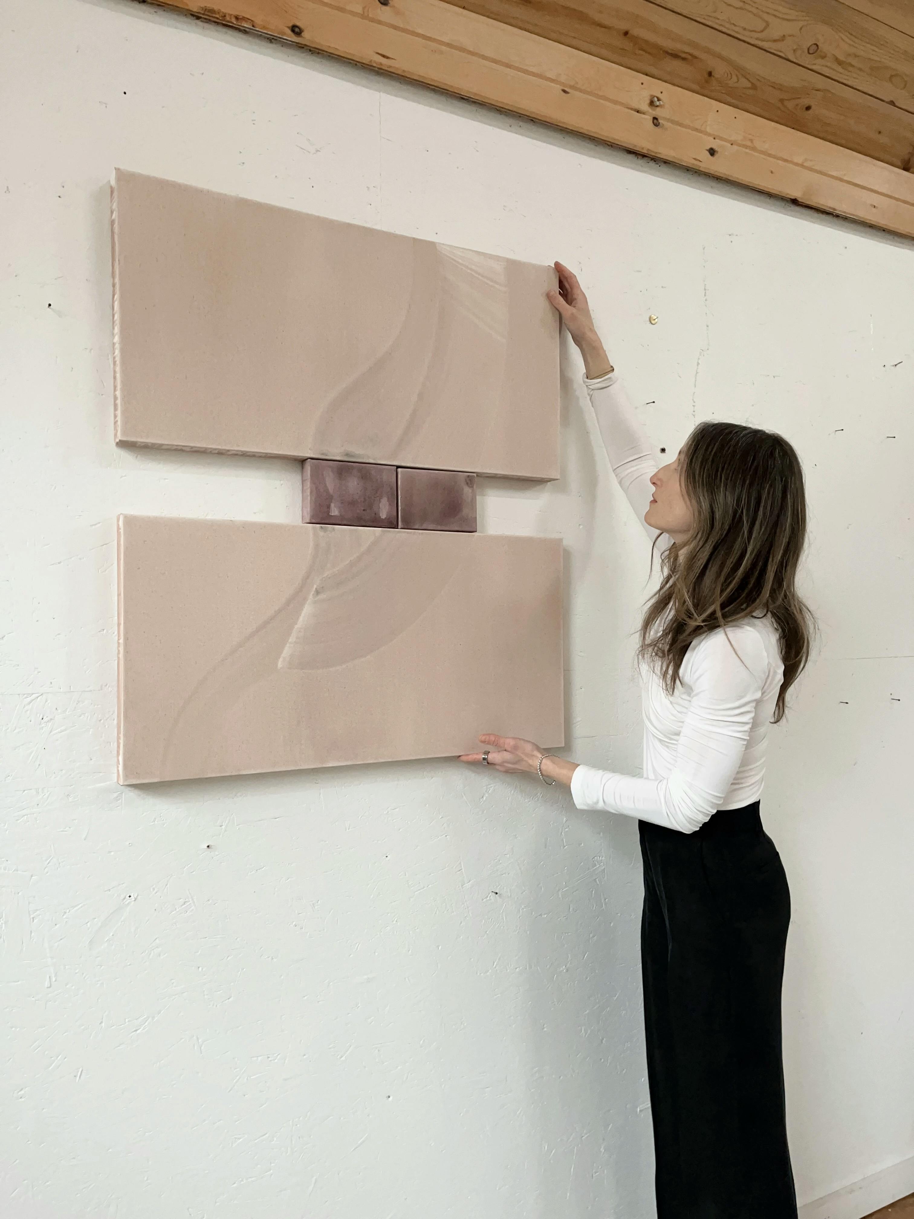 Artist Laura Naples holding up a pink, acrylic wash diptych against a white wall in her studio.